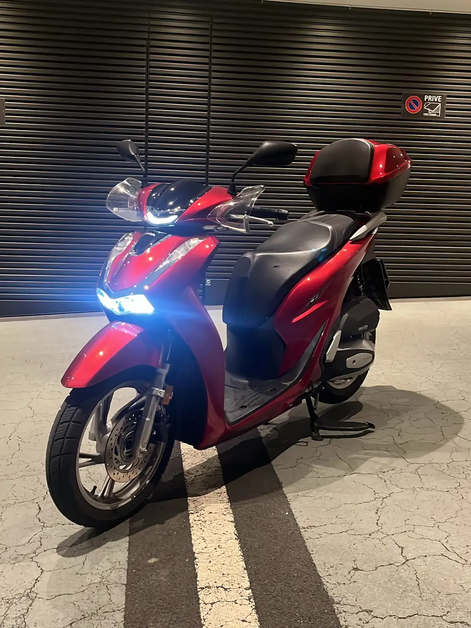 Roter Honda SH125i mit Topcase und eingeschaltetem Scheinwerfer in einem überdachten Parkplatz
