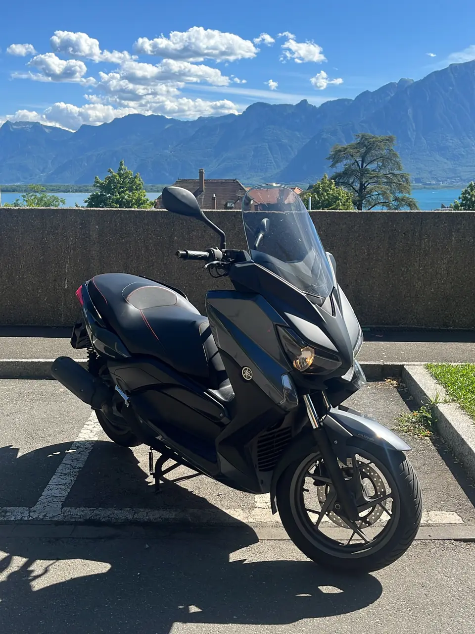 Schwarzer Yamaha XMAX 125 in der Sonne, Berge und See im Hintergrund